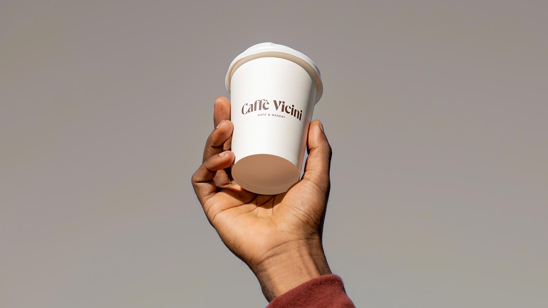 Hand holding a white Caffe Vicini Café & Bakery takeaway coffee cup against a clean, neutral background