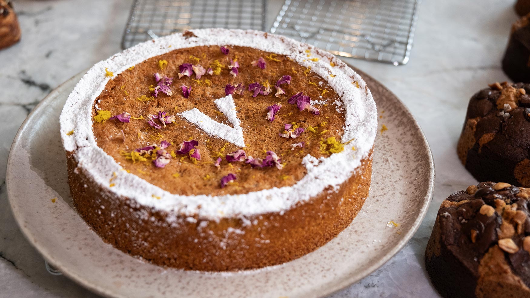 Round Caffè Vicini cake dusted with icing sugar, topped with edible flowers and a bold V design on a ceramic plate.
