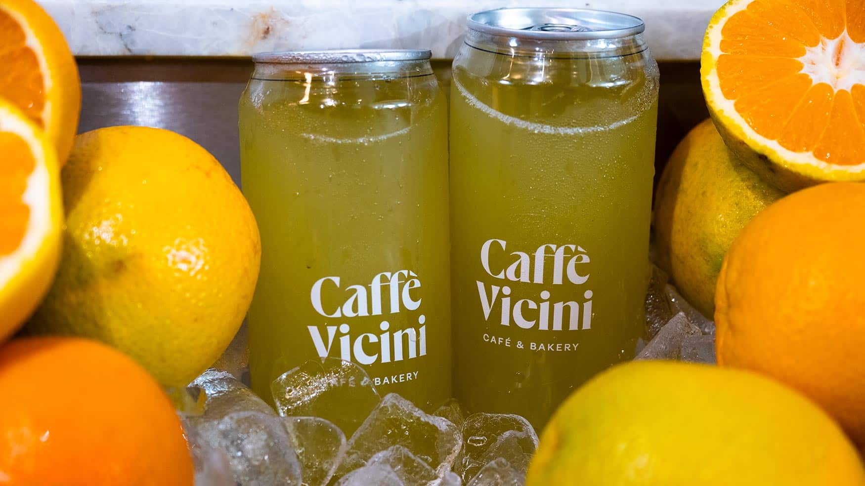 Two chilled cans of Caffè Vicini citrus drink on ice, surrounded by fresh oranges and lemons for a vibrant, refreshing display.