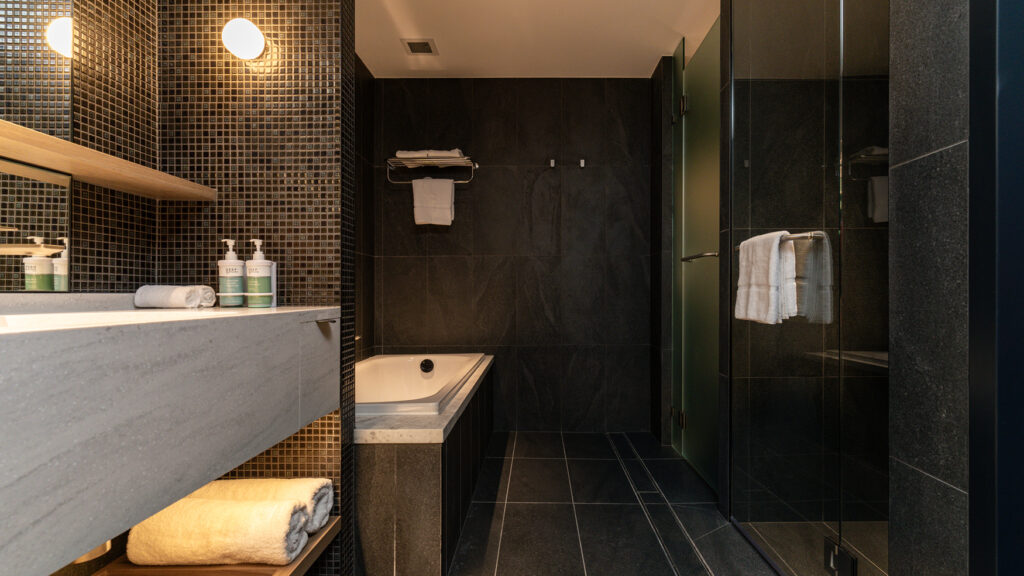 Black-tiled Presidential Suite bathroom with a bathtub, vanity and glass shower at Novotel Sydney Cabramatta