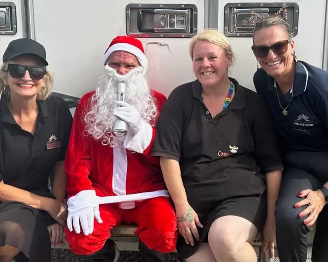 Cabravale Club Resort staff and Community Cafe volunteers sit with Santa Claus at a community Christmas event, smiling and celebrating with families and children.