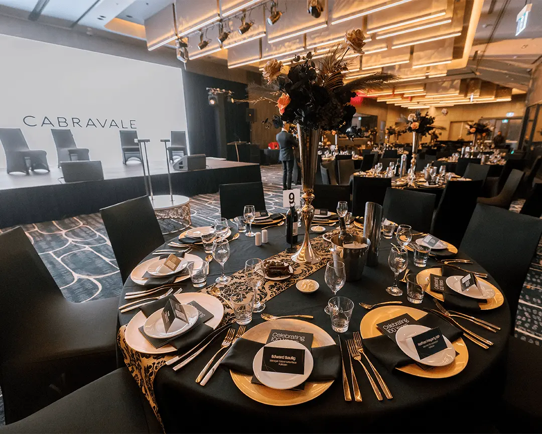 Formal gala dinner setup with round table, black tablecloth, gold floral centrepiece and Cabravale stage backdrop at the function venue