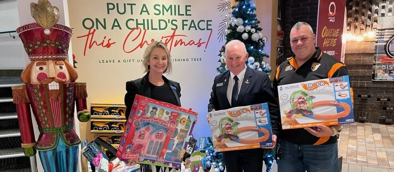 Cabravale Club Resort representatives hold donated toys beside a Christmas tree and sign reading “Put a Smile on a Child’s Face this Christmas!"