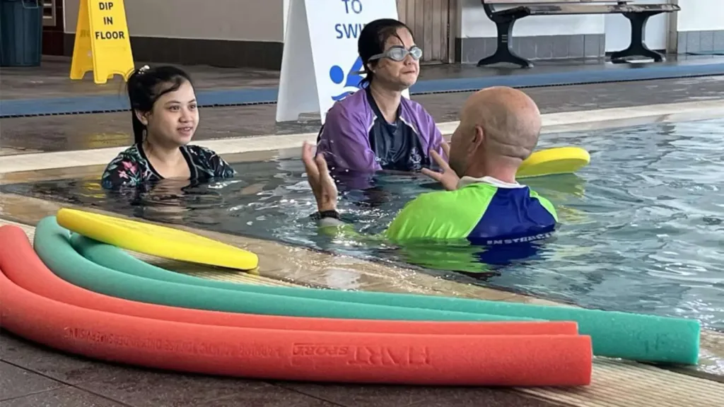 Swim instructor demonstrates techniques to adult learners during a CALD Learn to Swim class at a local indoor pool.