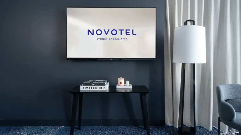 Wall-mounted TV displaying “NOVOTEL SYDNEY CABRAMATTA” above a console table and floor lamp in a modern hotel room interior
