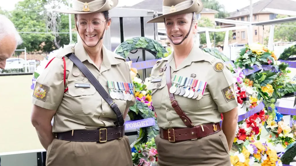 Two Australian Army servicewomen stand proudly in uniform with medals before wreaths reading “Lest We Forget”.