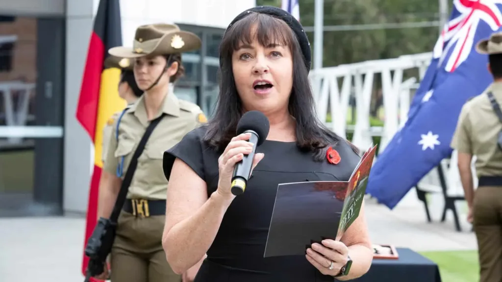A woman in black speaks into a microphone while holding a program, with uniformed service members and flags behind her.