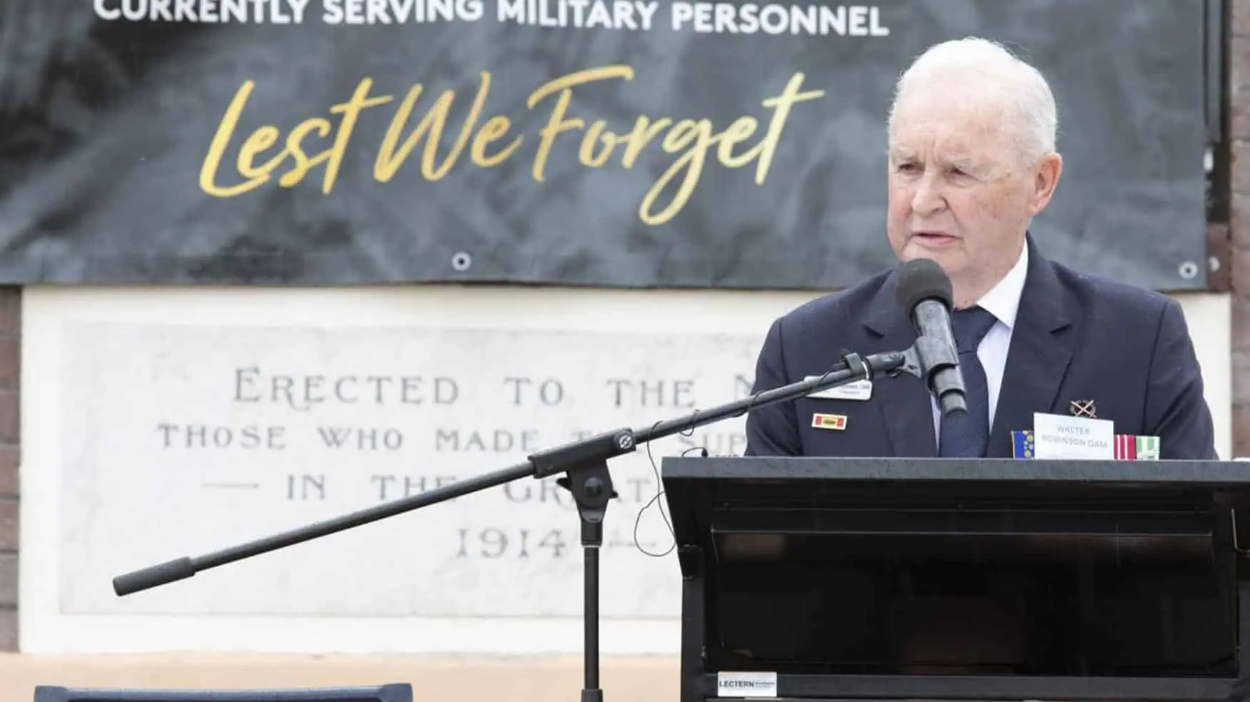 Veteran delivering a speech at a memorial podium during the Victory in the Pacific 2024 commemorative service at Cabravale, with a “Lest We Forget” banner behind him.