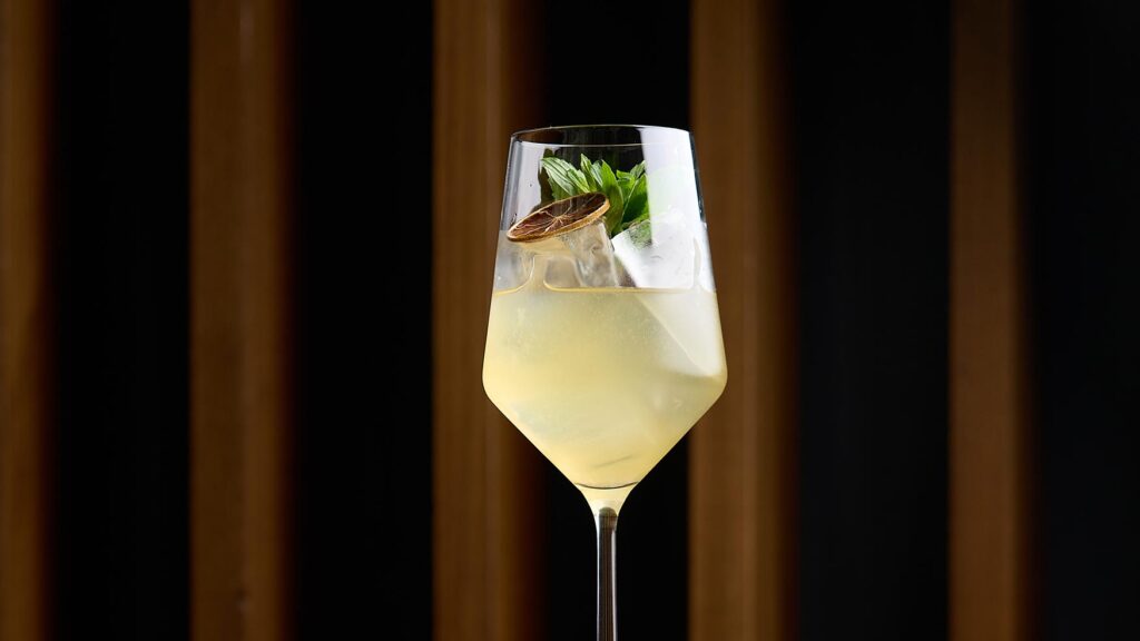 Tall glass with pale citrus cocktail garnished with dehydrated lime and herbs at Lotus Bar.