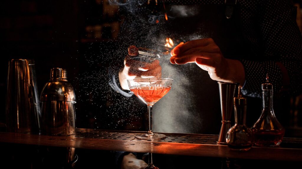 Bartender zesting citrus flame over a ruby cocktail, mist sparkling above the coupe in moody bar lighting at Lotus Bar.