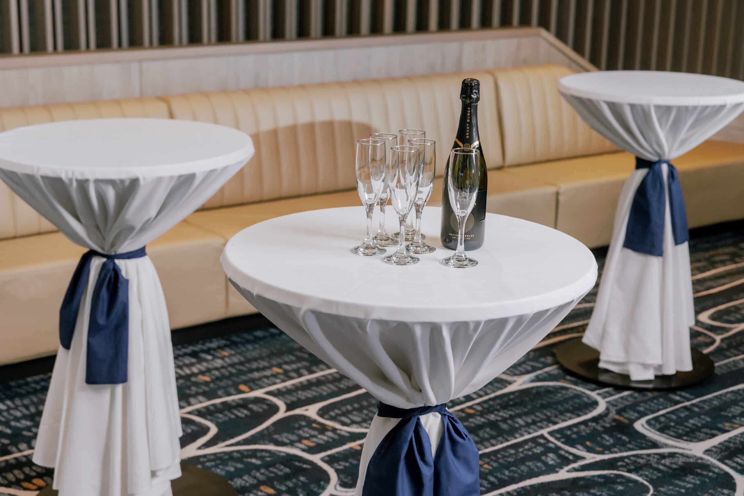 Styled cocktail tables draped in white linen with navy sashes, set with champagne bottle and glasses for an elegant event setting at Cabravale Club Resort