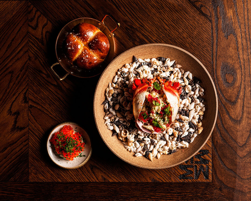 Overhead view of a plated seafood dish with a bread roll and relish on a timber table, part of the Stay & Dine package