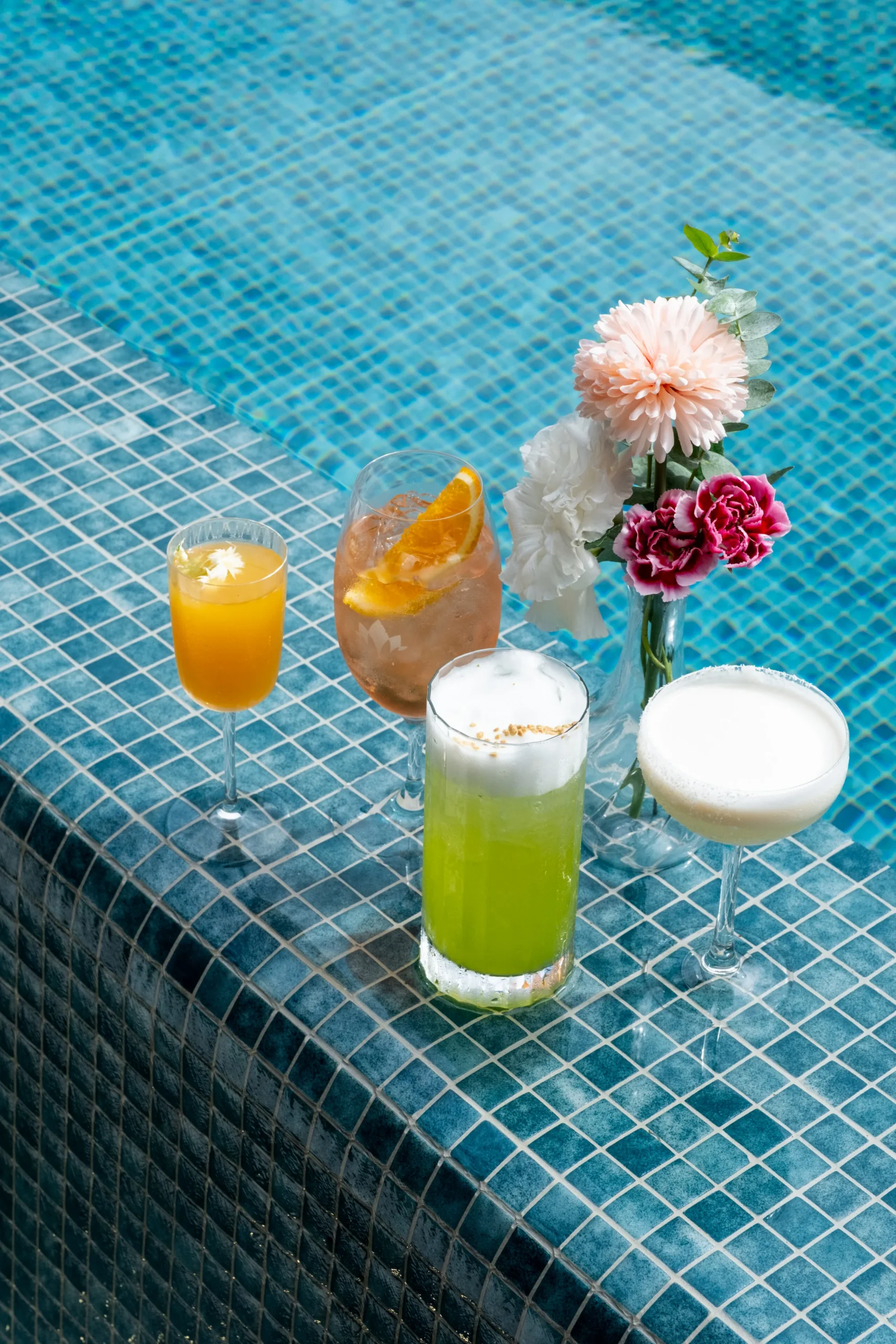 Four Mother's Day cocktails arranged on poolside tiles beside pink and white floral bouquet