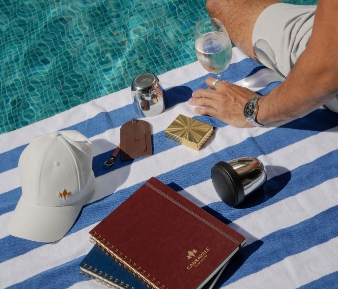 Cabravale branded notebook, cap and leather card holder styled poolside on a striped towel