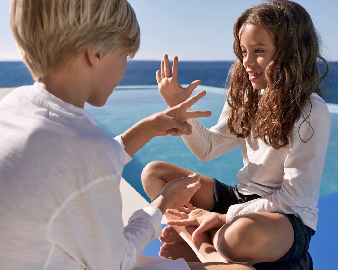 Two children playing rock paper scissors by an infinity pool with ocean view in the background