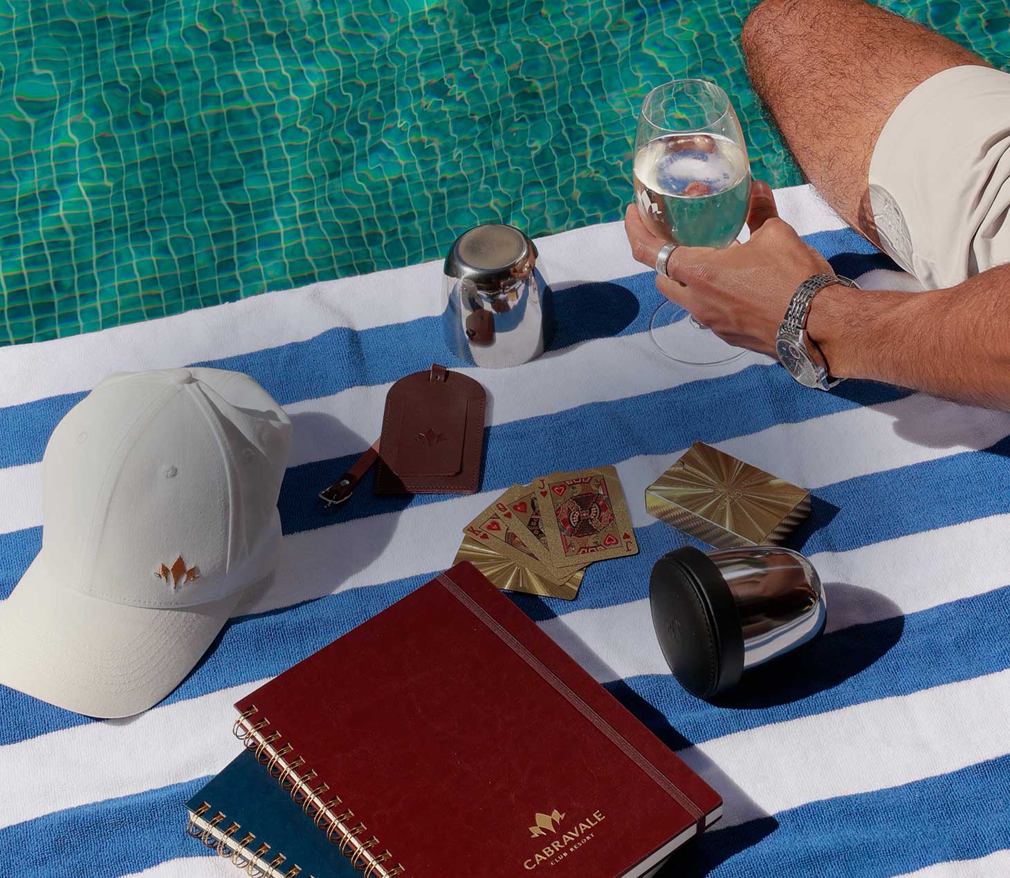 White Cabravale Boutique cap, red branded notebook and gold playing cards styled on a striped poolside towel