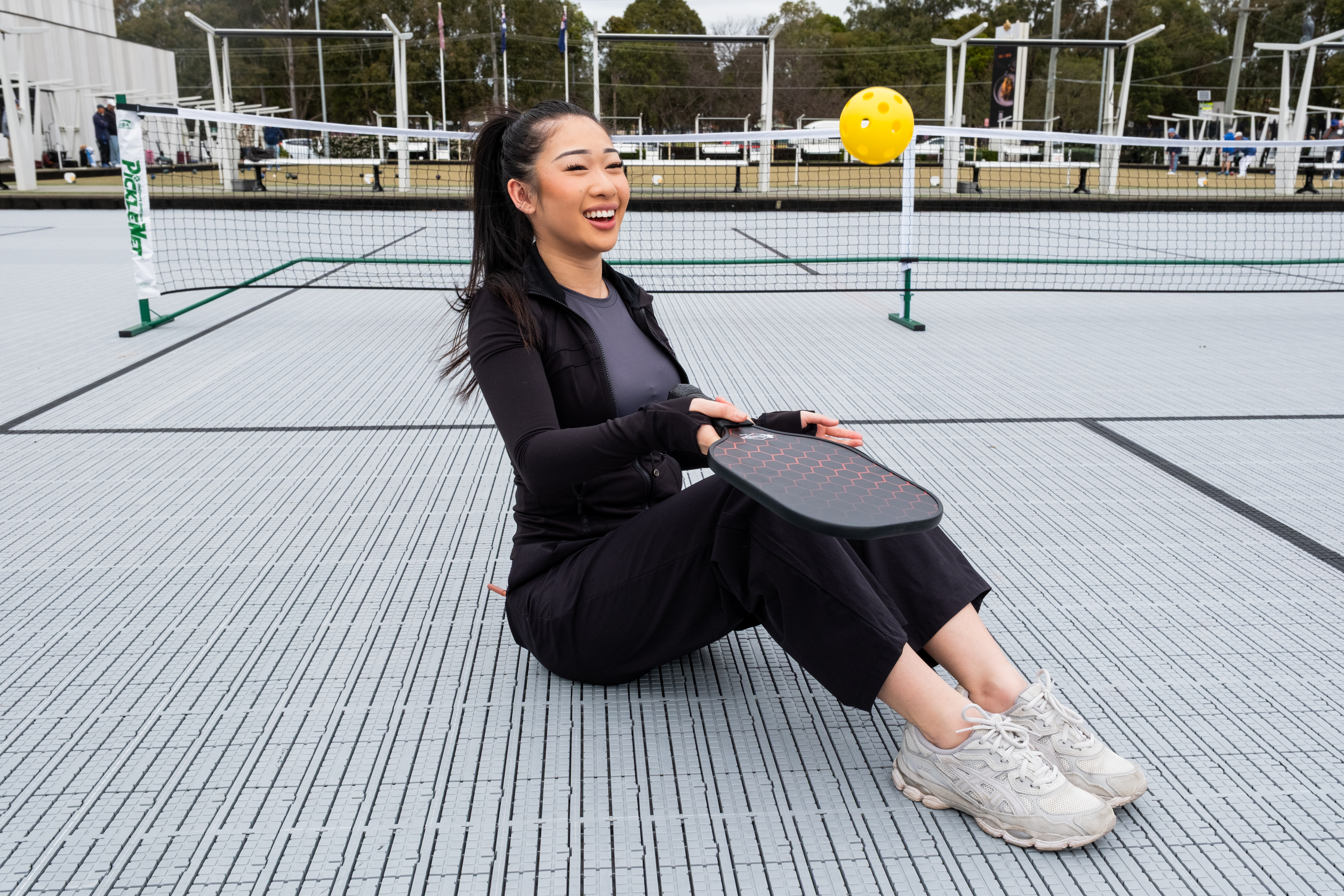 Girl sitting on pickleball court laughing while hitting a ball