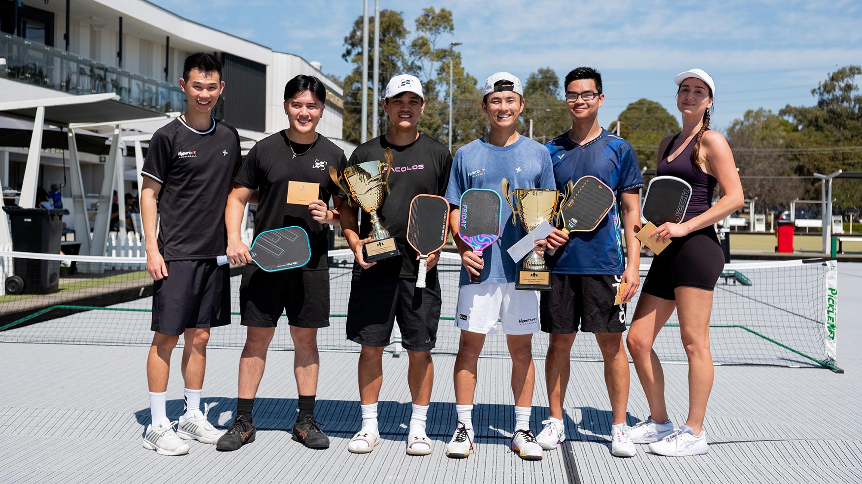Six pickleball players holding trophies and paddles on the Cabravale pickleball courts in Cabramatta