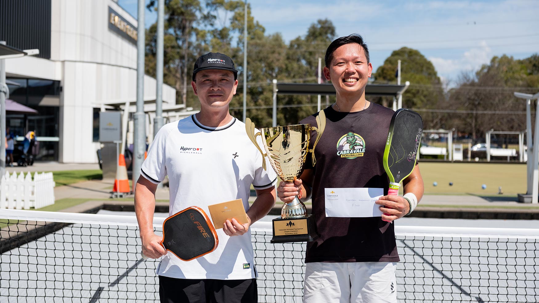 Two tournament winners holding a trophy, paddles and a prize envelope at Cabravale Pickleball in Cabramatta
