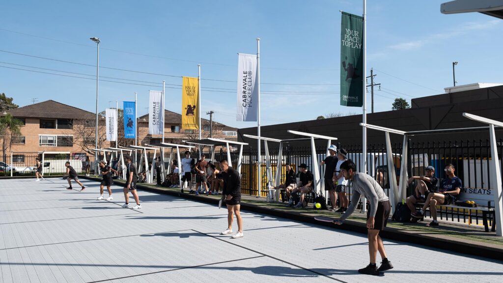 Players and spectators across the outdoor pickleball courts in Cabramatta, with shaded seating along the fence